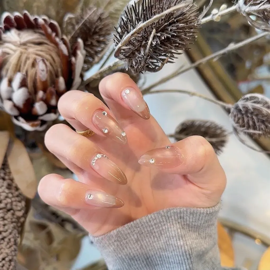 Long almond nails in sheer nude with a golden chrome shimmer and crystal gems